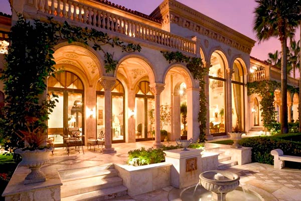 Exterior of a Mediterranean-style estate at twilight, with arched colonnades, illuminated interiors, palm trees, and a fountain in the foreground.